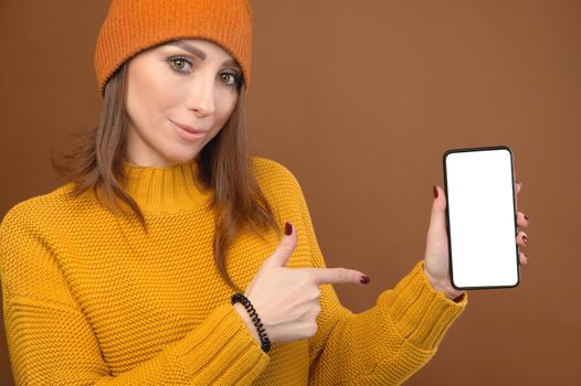 Smiling caucasian young lady pointing with finger at smartphone in hand, standing against orange studio background