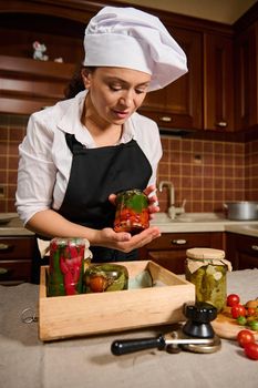 Beautiful woman, housewife puts a jar of fermented cherry tomatoes upside down on a wooden crate with canned vegetables