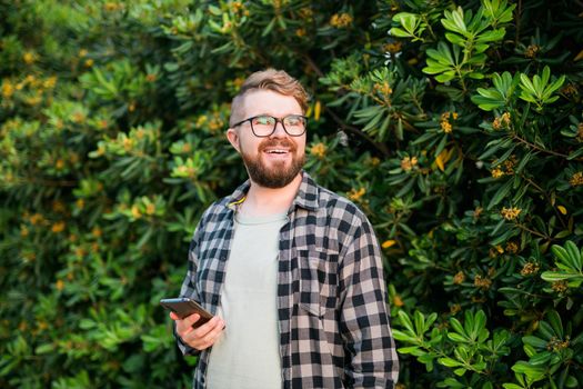 Portrait of attractive cheerful guy using smartphone for scrolling on social media over green tree background copy space and empty place for text