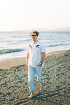 Young man with glasses walks barefoot on the beach