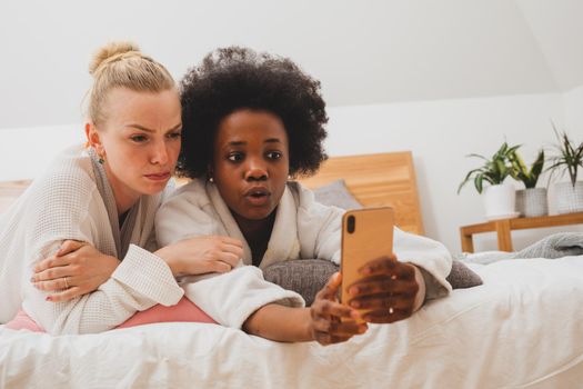 Two young women of different nationalities have a video chat at the spa. The women in bathrobes are lying on the bed and communicating to using a smartphone