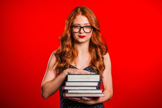 Serious female student on red background in studio holds stack of university books from library. Woman in glasses.