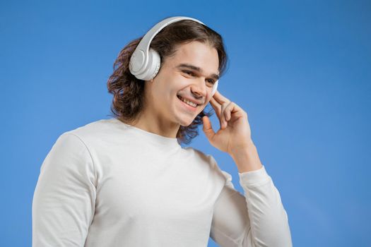 Charming young man with long hairdo listening to music with wireless headphones, guy having fun, smiling in studio on blue background. Dance, radio concept.