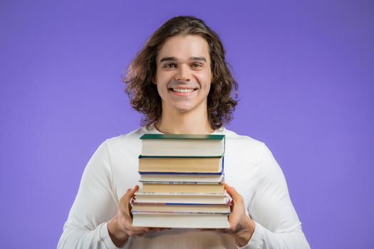Student holds stack of university books from library on violet background in studio. Man smiles, he is happy to graduate.