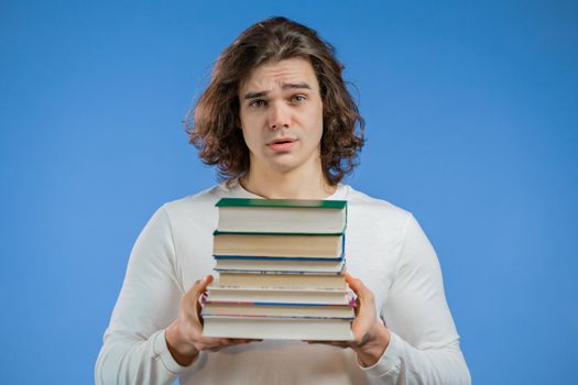European student is dissatisfied with amount of homework and books on blue background. Man with long hairdo in displeasure, he is annoyed,discouraged frustrated by studies