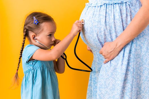 Motherhood, hot summer - croped portrait pregnant mother unrecognizable woman blue dress little Daughter girl sibling treat mom role play game stethoscope unborn big belly tummy on yellow background
