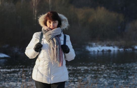 winter walk in nature. Cold season. Woman in white jacket with scarf and black gloves standing near river in forest.