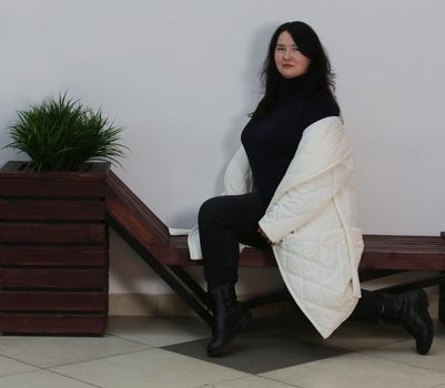 Cute plus-size brunette sitting on a bench in a white coat.