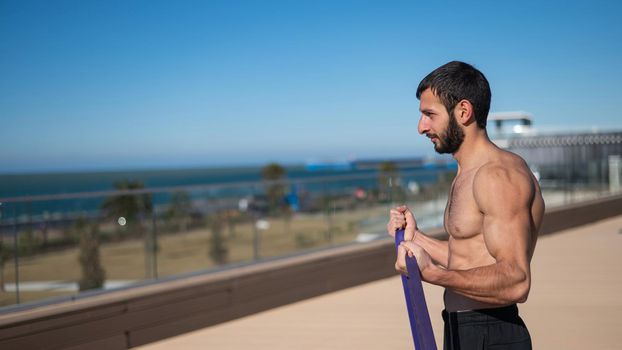 Shirtless man doing exercise with rubber bands outdoors.