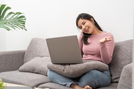 Happy young asian woman calling with mobile phone and usin laptop computer on sofa at home