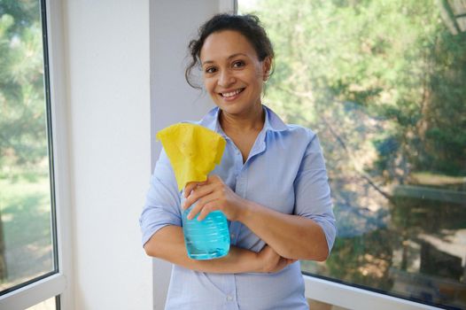 Pleasant housewife smiles stands against panoramic veranda windows with glass cleaner detergent and yellow rag in hands