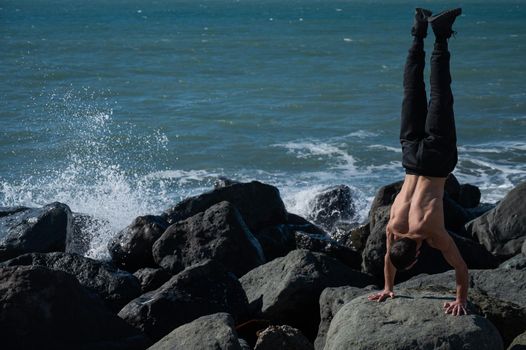 Shirtless man doing handstand on rocks by the sea.