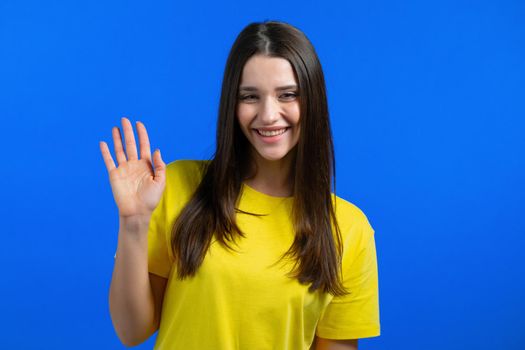 Friendly woman waving hand - goodbye, chao, adios. Parting, say bye to camera. Beautiful girl waving hand on blue studio background