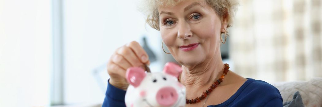 Elderly woman putting coin into piggy bank at home