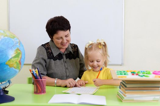 Mature teacher sitting at table teaching little girl how to write