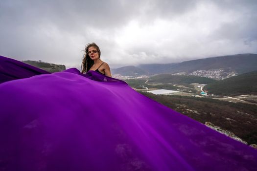 A woman with long hair is standing in a purple flowing dress with a flowing fabric. On the mountain against the background of the sky with clouds.