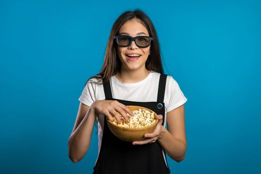 Young asian girl in 3d glasses watching fascinating comedy movie, laughing and eating popcorn on blue studio background.