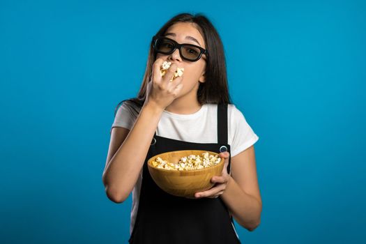 Young asian girl in 3d glasses watching fascinating comedy movie, laughing and eating popcorn on blue studio background.