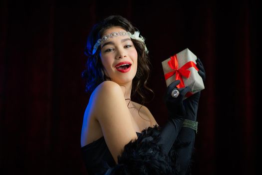 Joyful vintage styled woman dressed in Roaring twenties era holding gift box with bow on velours background. She is glad to get present. retro, party, fashion concept.