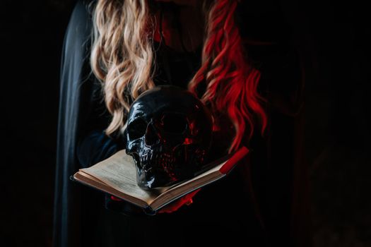 Witch in black costume with black magic book. Woman holds skull in hands. Death, spiritual rituals concept, Halloween, horror, scary symbol of dead.
