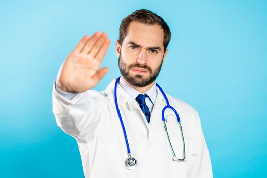 Portrait of serious doctor in professional medical white coat showing rejecting gesture by stop palm sign. Doc man isolated on blue background.