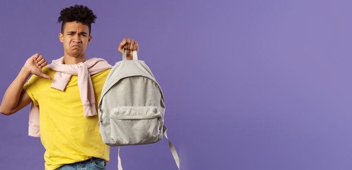 Education, university and trends concept. Portrait of gloomy disappointed young male student complaining on ugly new backpack, show thumbs-down and grimacing displeased, purple background