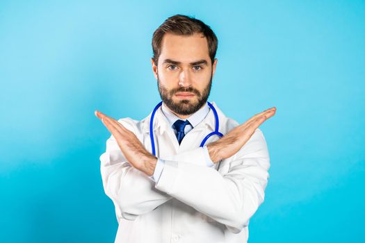Portrait of serious doctor in professional medical white coat with no crossing hands sign make negation gesture. Denying, Rejecting, Disagree. Doc man isolated on blue background.