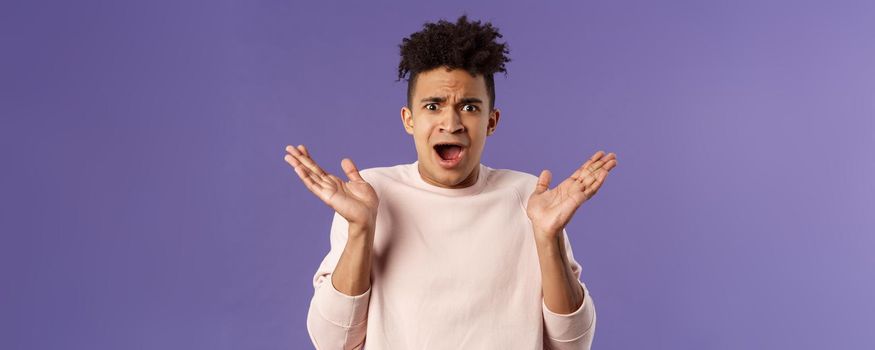 Close-up portrait of displeased, bothered frustrated hispanic man spread hands sideways in dismay and confusion, staring camera upset with disappointed grimacing expression, purple background