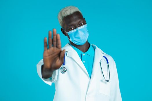 Portrait of serious black doctor in professional medical white coat and mask showing rejecting gesture by stop palm sign. Doc man isolated on blue background.