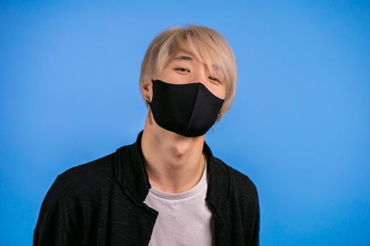Portrait of young asian man in black protective mask. Hipster guy with trendy dyed blond hairdo and earrings. Blue studio background.