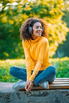 African teenager girl listens to music with headphones in park. Cute woman in yellow, dancing with head, curly hair. Student, freedom, music concept.