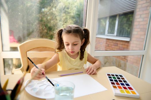 Top view. Preschooler girl in a yellow T-shirt and two ponytails, enjoying watercolor painting during art class at home