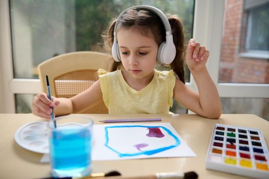 Beautiful European little girl, child of preschool wearing wireless headphones is painting with watercolors in classroom