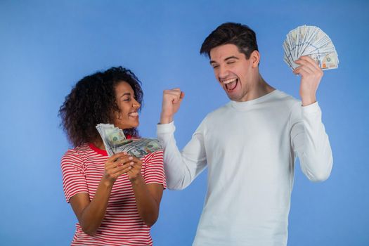 Amazed happy excited interracial couple showing money - USD currency dollars banknotes on blue wall. Symbol of success, gain, victory. African woman and white man win lottery.