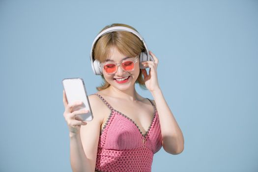 Photo of excited woman in swimwear isolated over blue wall background showing display of mobile phone listening music with headphones.