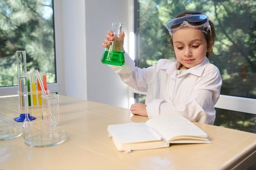 Delighted schoolgirl in white lab coat and safety goggles, testing chemistry lab experiment and shaking glass tube flask