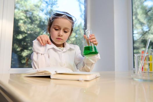 Thoughtful Caucasian little girl in safety goggles and white lab coat, concluding science experiments in chemistry class