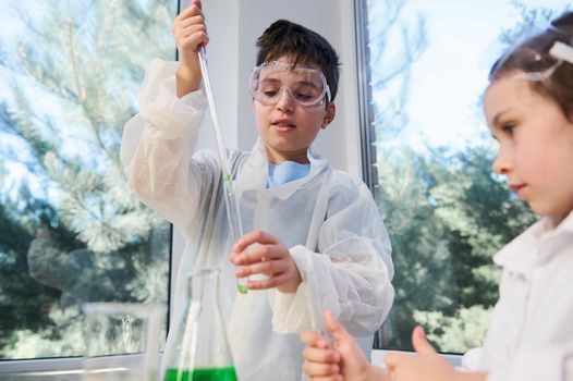 Handsome schoolchild in lab safety wear using pipette, dripping reagent into a test tube, making experiment at chemistry