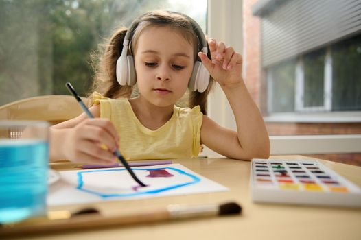 European preschooler girl in wireless headphones, listening to the music while painting and drawing a picture on paper.