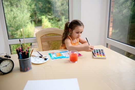 Caucasian child, elementary age girl sitting at a table and painting picture with watercolors and paintbrush.