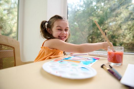Cheerful little preschooler girl, smiles looking at the camera while painting picture with watercolors and paintbrush.