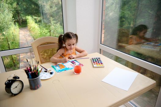 Top view of adorable child, elementary age girl sitting at a table and painting picture with watercolors and paintbrush.