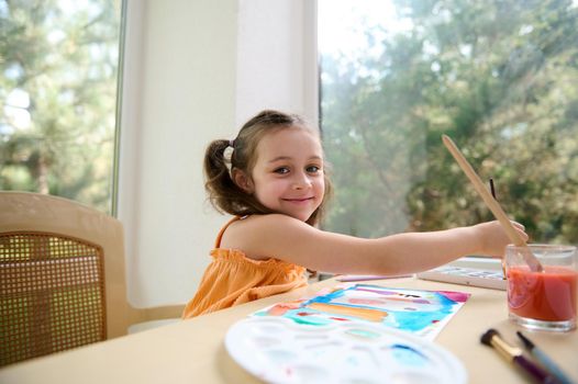 Adorable little preschooler girl, smiles looking at the camera while painting picture with watercolors and paintbrush.