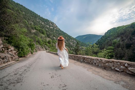 Female traveler in brown hat and white dress looking at amazing mountains and forest, wanderlust travel concept, atmospheric epic moment