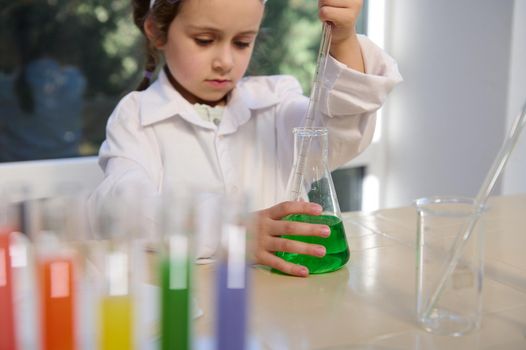 Focus on flat-bottomed flask with solution and little girl's hand using pipette, collects chemicals, doing experiments