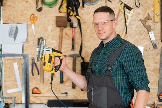 Portrait of a man with an electric jigsaw on wood in the workshop.