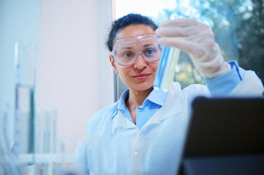 Close-up of beautiful African American woman in safety goggles examining test tubes with chemical substances, in biolab