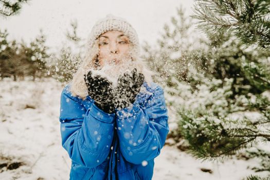 Young cute girl blowing on first snow from hands to camera and then scatters it to sides. Winter forest background. Portrait of cute woman childishly rejoices nature.