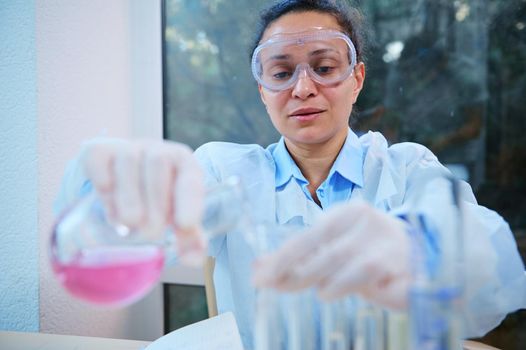 African American woman, chemist pharmacologist pours pink fluid substance from a glass laboratory flask into test tubes