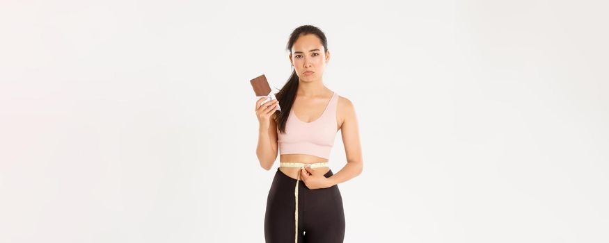 Sport, wellbeing and active lifestyle concept. Disappointed gloomy asian girl measuring waist with tape measure and sulking as cannot eat chocolate bar while losing weight on diet